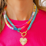 Girl wearing three necklaces: a big blue beaded necklace with a gold heart shape medallion attached to it, a multicolored beaded short necklace, and a thin multicolored beaded choker