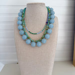 Three necklaces on a mannequin bust against a white background. Chunky blue bead necklace, green necklace and dainty blue bead necklace