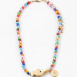 Picture of a bright multi-colored bead necklace with a tan fish bead and a gold disc pendant.