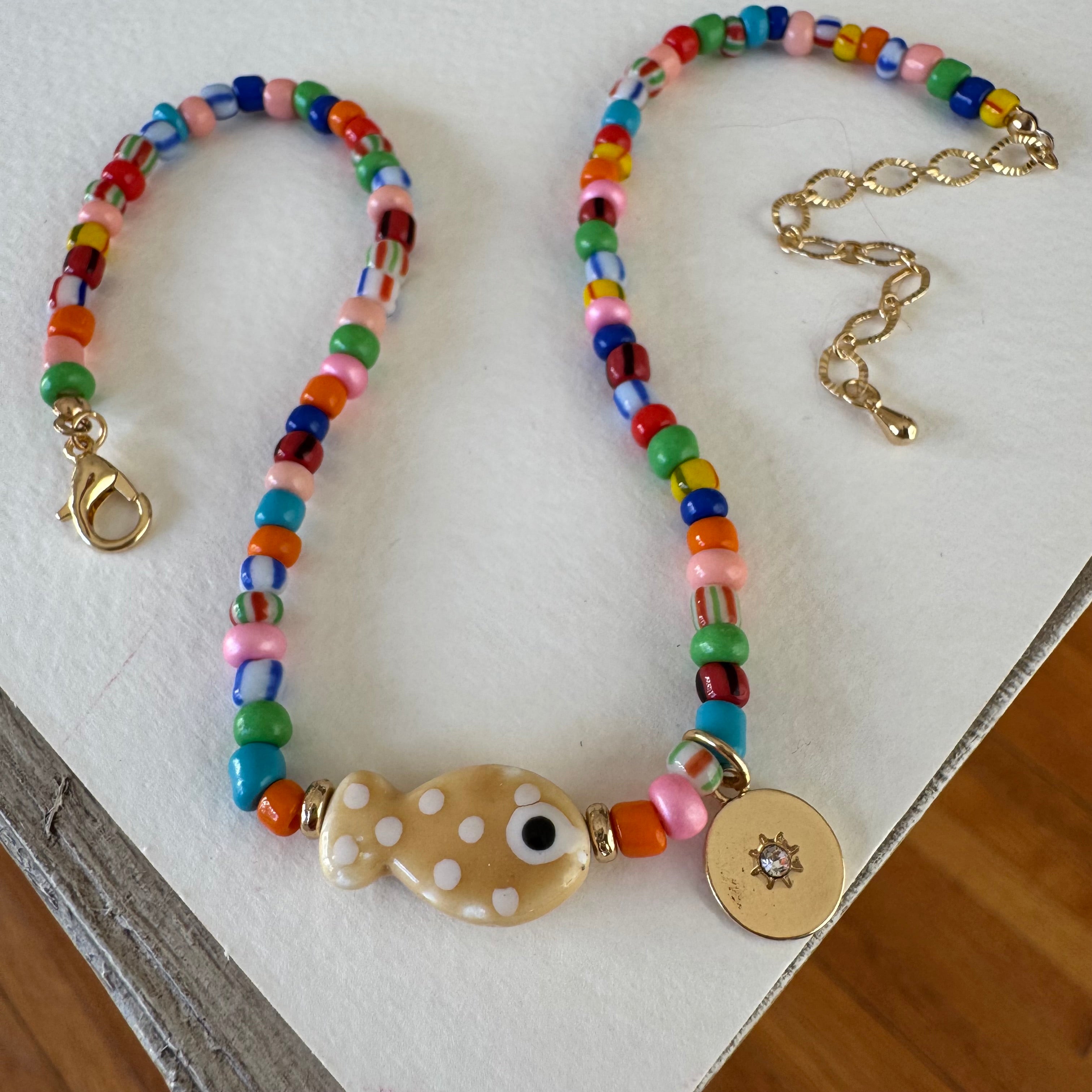 Picture of a multicolor bead necklace with a fish pendant at its center and a disc pendant with a diamond in the center.