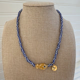 Bead necklace made of white and black striped beads with a tan polka dot fish bead at the center and a small gold disc pendant.