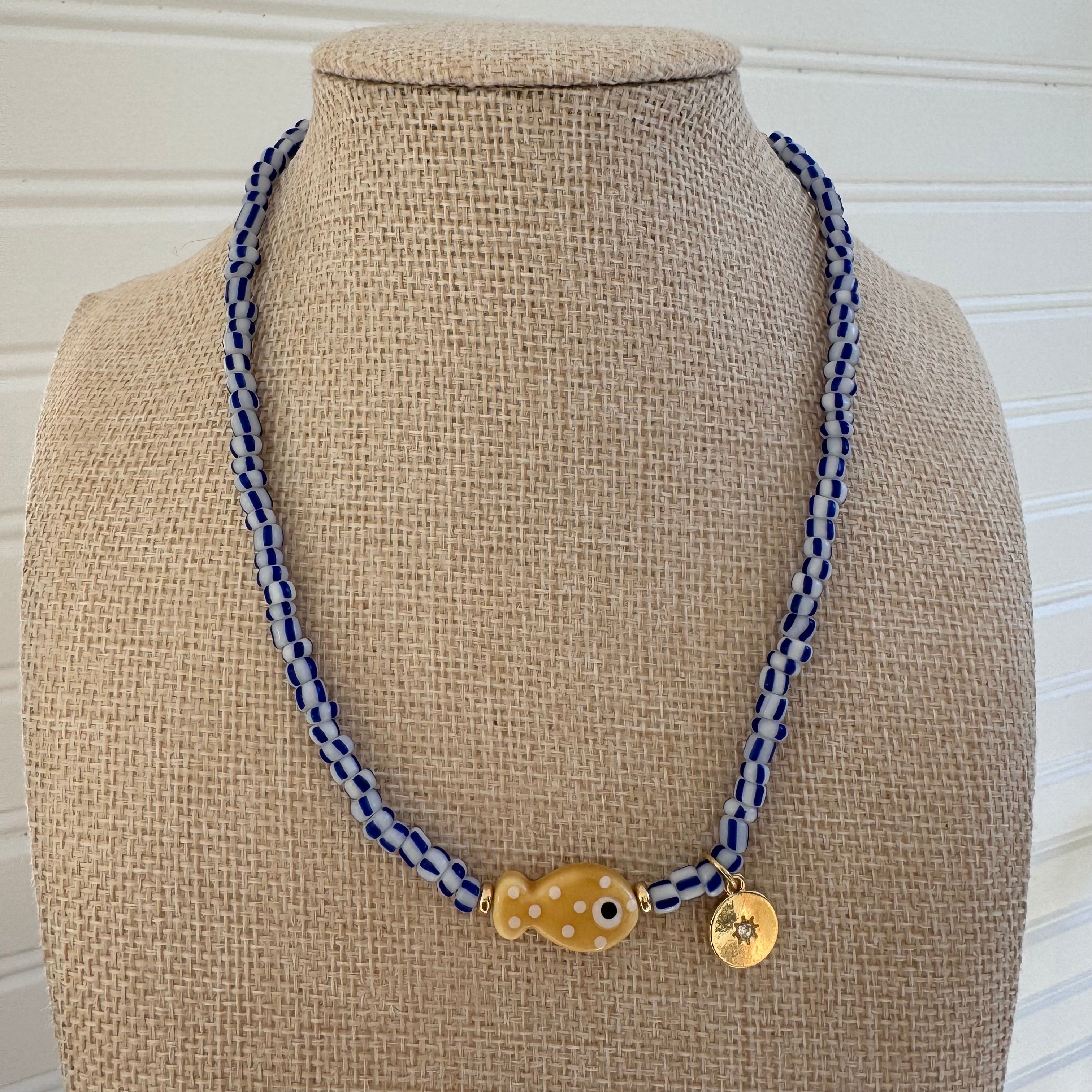 Bead necklace made of white and black striped beads with a tan polka dot fish bead at the center and a small gold disc pendant.