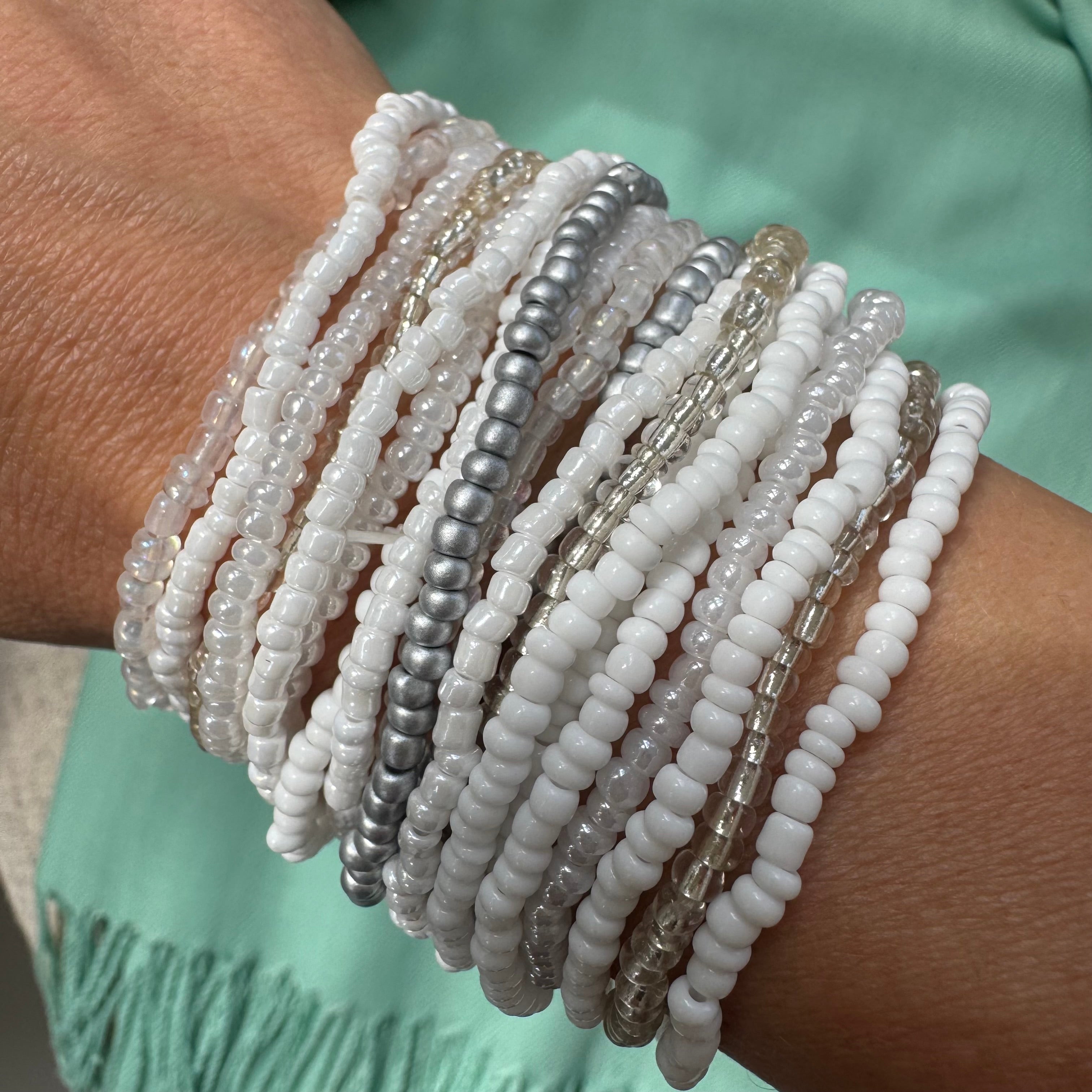 Multiple white and pearlescent beaded bracelets on a wrist with a blurred green background
