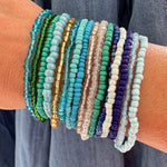 Multiple beaded bracelets of various blue, green and beige colors on a wrist against a denim background
