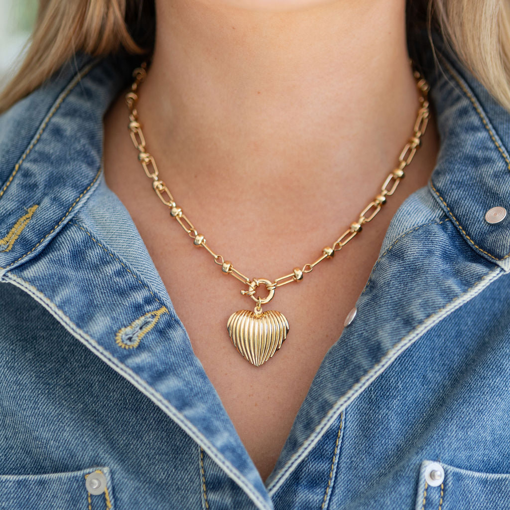 Gold heart-shaped pendant on a bold gold chain necklace worn by a person in a denim jacket.