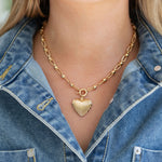 Gold heart-shaped pendant on a bold gold chain necklace worn by a person in a denim jacket.