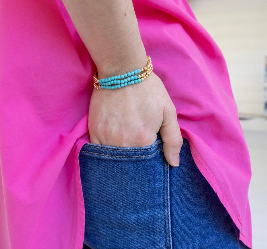 Person wearing a turquoise and gold beaded bracelet on a pink fabric background