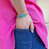 Person wearing a turquoise and gold beaded bracelet on a pink fabric background