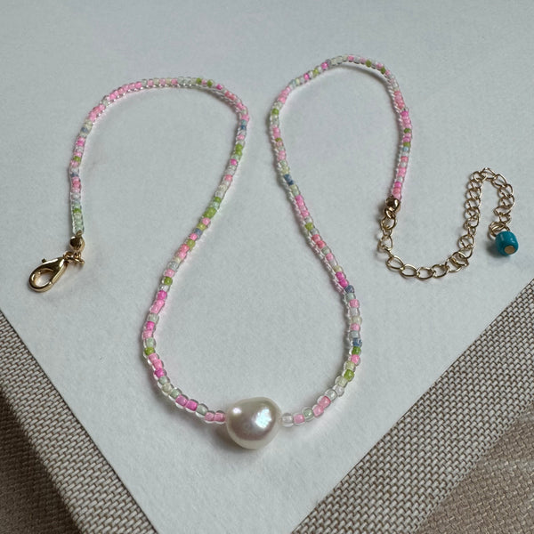Picture of a delicate seed bead necklace with clear pink, greens and white beads. Center of necklace is a large freshwater Pearl.