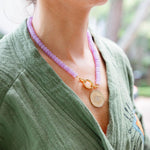 A girl wearing a light pink stone beaded necklace with a gold chain link in the middle with a gold engraved eye medallion attached to it
