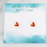 A pair of red heart-shaped stud earrings with gold trim, displayed on a blue watercolor background card with the text 'well aren't you looking pretty today!'.
