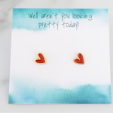 A pair of red heart-shaped stud earrings with gold trim, displayed on a blue watercolor background card with the text 'well aren't you looking pretty today!'.