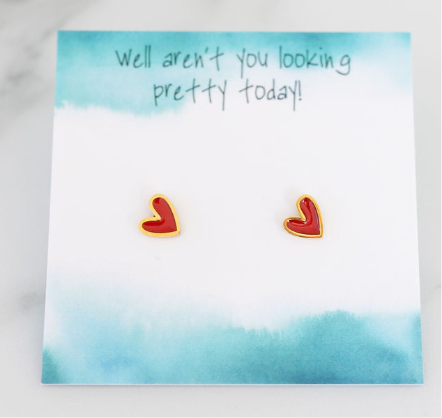 A pair of red heart-shaped stud earrings with gold trim, displayed on a blue watercolor background card with the text 'well aren't you looking pretty today!'.