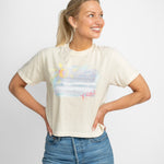 Happy blonde woman wearing a cream colored t shirt tucked into her blue jeans. T shirt has a beach scene printed on the front of it with the letters XOXO in pink.