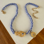 A bead necklace made of blue and white striped beads with a tan polka dot fish bead at its center and a small gold disc pendant.