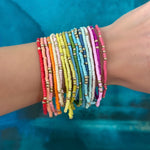 Picture of a wrist wearing 19 different colored seed bead bracelets with accent gold beads.  Bracelets are red, orange, beige, pinks, white, yellows, blues, greens and purple.