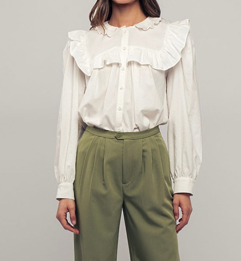 Picture of a model wearing a white long sleeved dress shirt. Shirt has a ruffle collar around the neck and breast and is a button up shirt.