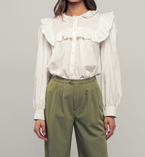 Picture of a model wearing a white long sleeved dress shirt. Shirt has a ruffle collar around the neck and breast and is a button up shirt.