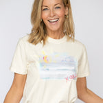 Happy blonde woman wearing a cream colored t shirt tucked into her blue jeans. T shirt has a beach scene printed on the front of it with the letters XOXO in pink.