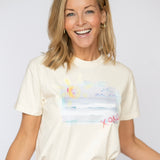 Happy blonde woman wearing a cream colored t shirt tucked into her blue jeans. T shirt has a beach scene printed on the front of it with the letters XOXO in pink.