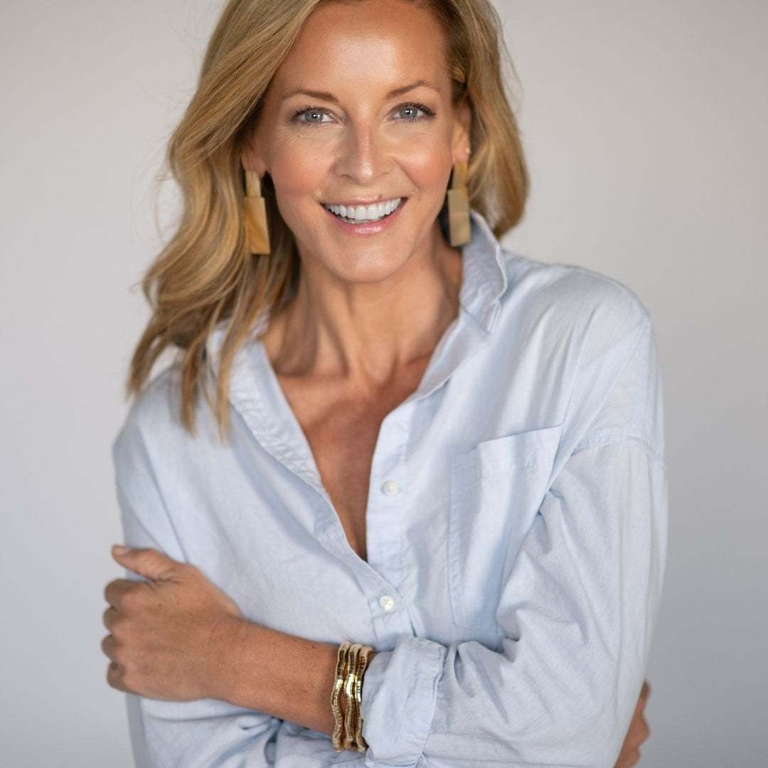 Woman wearing a light blue button up shirt with large gold statement earrings and bracelets on a plain background