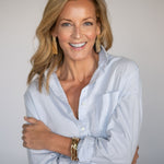 Woman wearing a light blue button up shirt with large gold statement earrings and bracelets on a plain background
