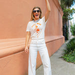 Woman wearing sunglasses and a cream shirt with an orange print of a cowgirl with a lasso and the text "Stay Wild" with multiple blue beaded necklaces smiling and raising her arm in the air
