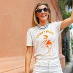 Woman wearing sunglasses and a cream shirt with an orange print of a cowgirl with a lasso and the text "Stay Wild" with multiple blue beaded necklaces smiling and raising her arm in the air