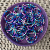 Picture of a large purple bowl filled with dozens of the same beaded bracelet. Bracelet displayed in shades of blue, purple, pink and some greens.