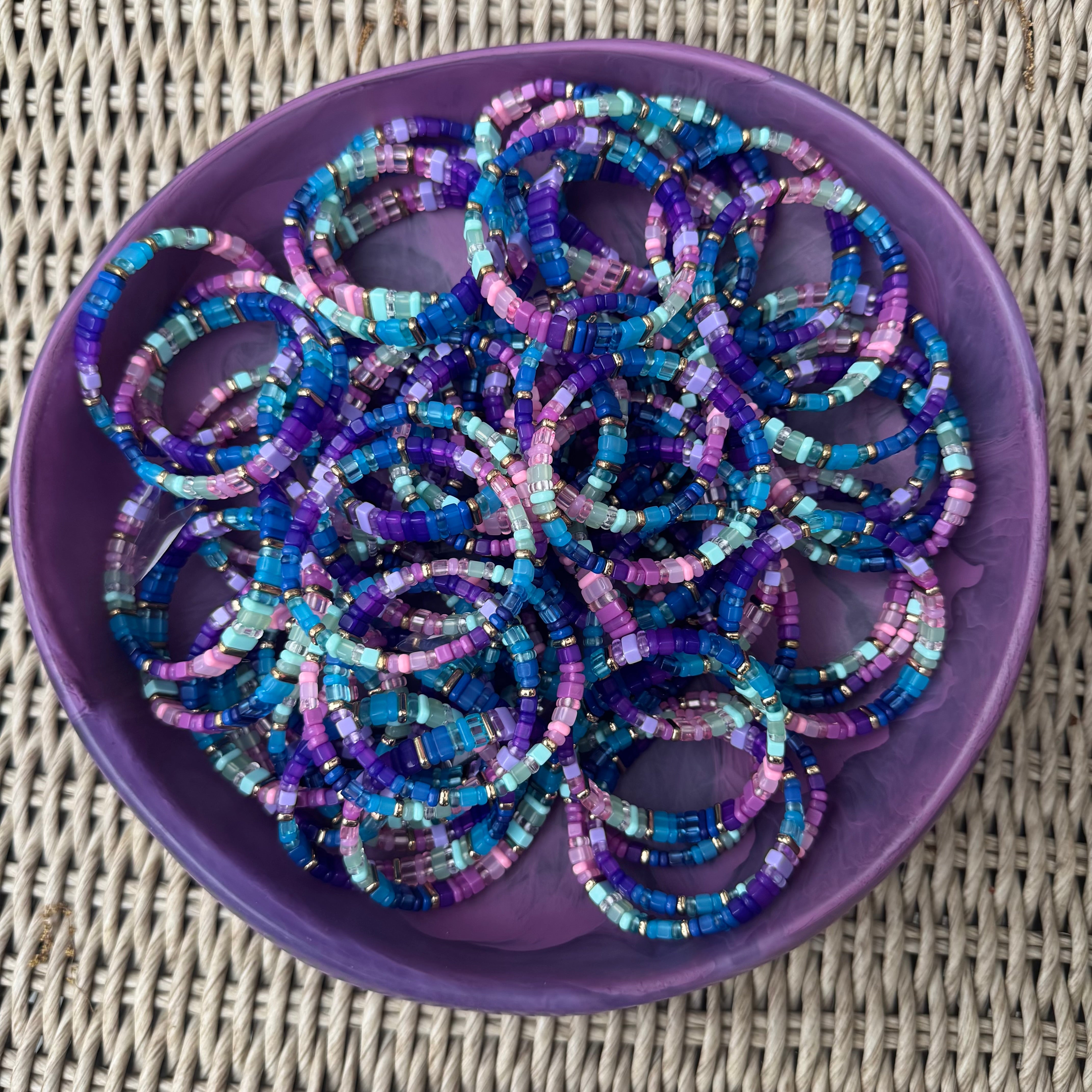 Picture of a large purple bowl filled with dozens of the same beaded bracelet. Bracelet displayed in shades of blue, purple, pink and some greens.