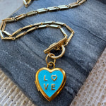 Gold chain necklace with a blue heart-shaped locket pendant on a textured surface