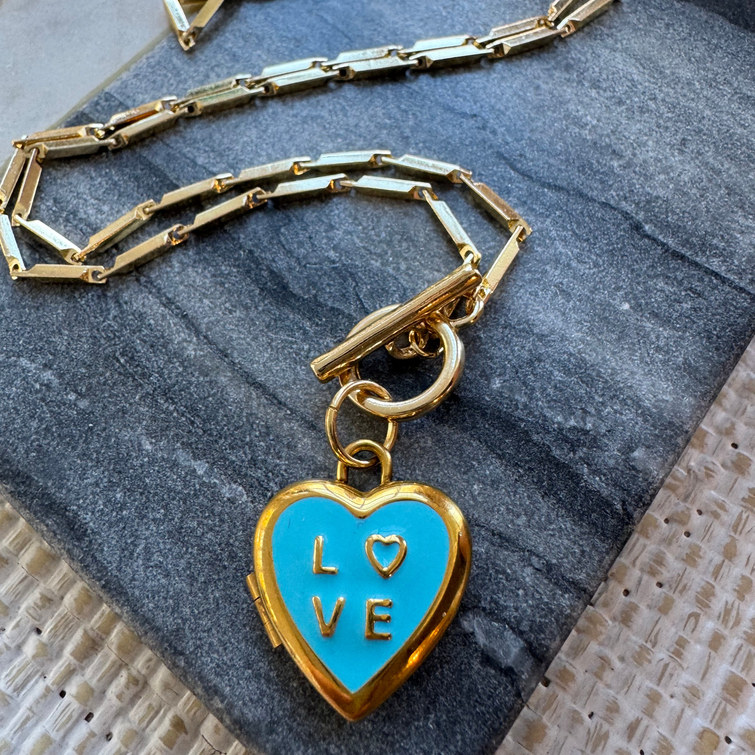 Gold chain necklace with a blue heart-shaped locket pendant on a textured surface
