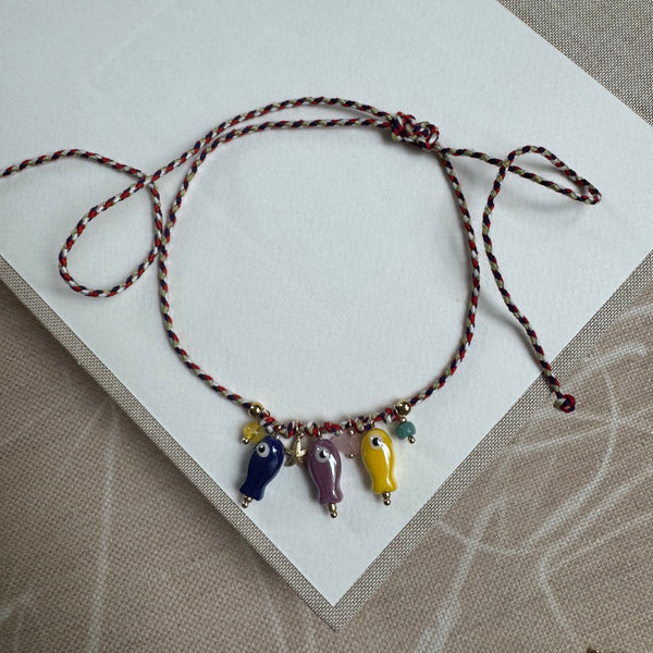 Picture of a thin woven  adjustable string bracelet in red, white and black. Dangling from it are three fish beads in blue, purple and yellow with small yellow, pink and green accent beads and a small gold starfish.
