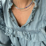 Picture of a woman wearing a blue blouse and two dainty bead necklaces. One is made of white and gold beads and one is of dark blue and light blue beads.