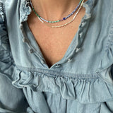 Picture of a woman wearing a blue blouse and two dainty bead necklaces. One is made of white and gold beads and one is of dark blue and light blue beads.