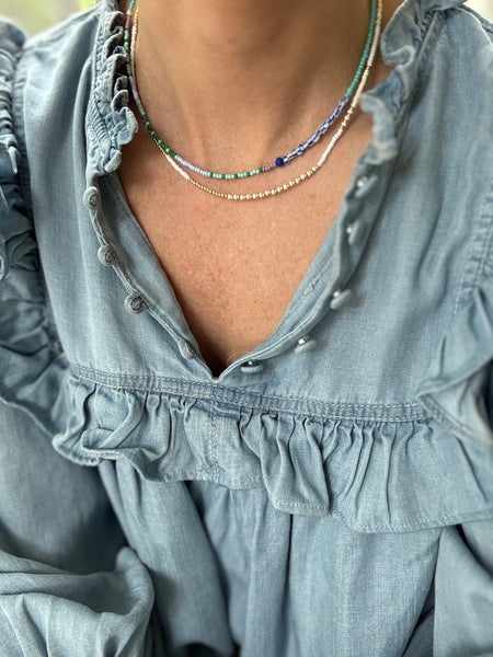 Picture of a woman wearing a blue blouse and two dainty bead necklaces. One is made of white and gold beads and one is of dark blue and light blue beads.