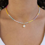 Dainty bead necklace in white, teal, yellow and pink with a small white enamel disc pendant.
