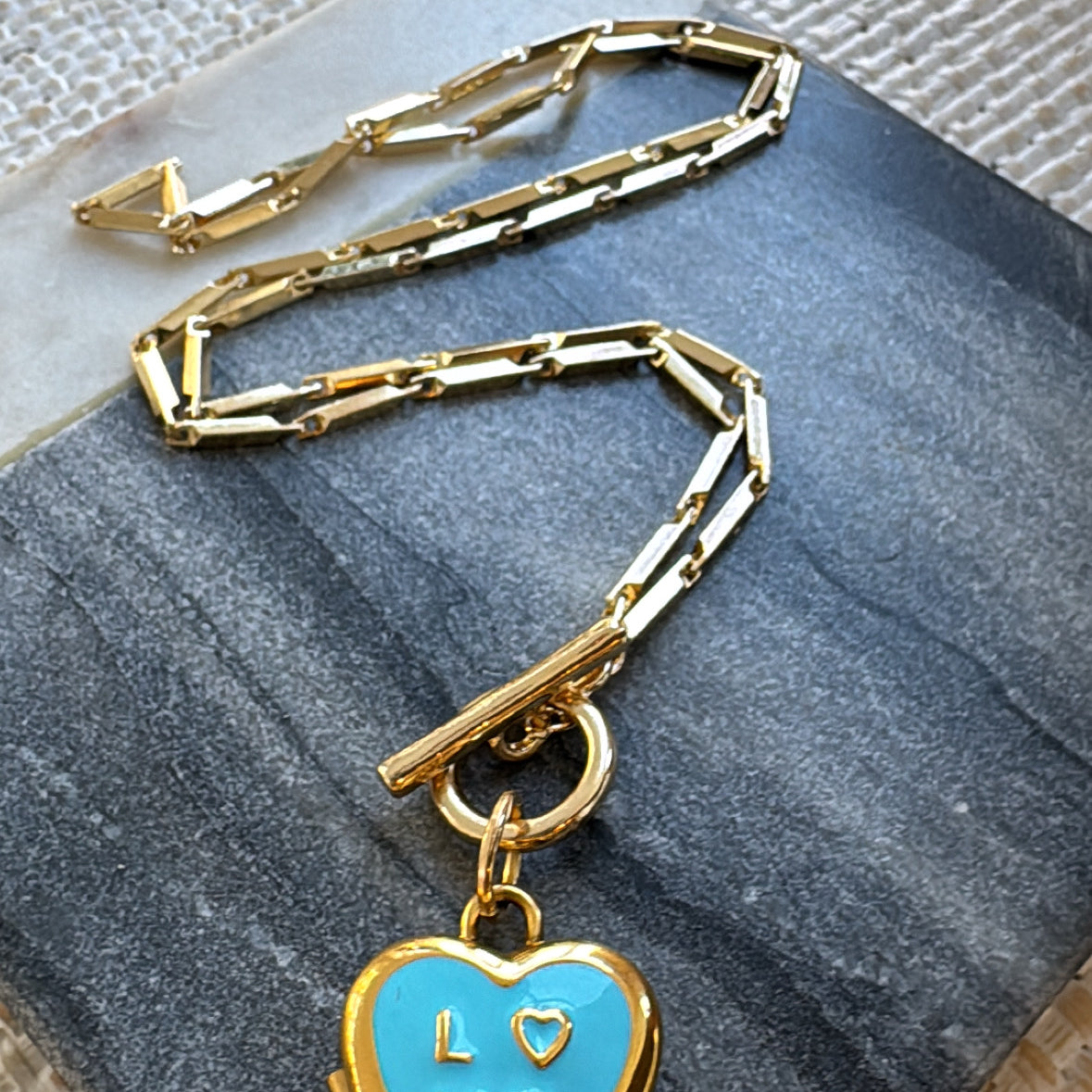 Gold chain necklace with a blue heart locket pendant on a textured surface