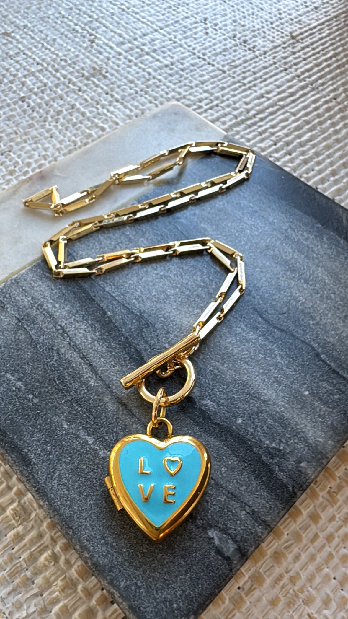 Gold chain necklace with a blue heart locket pendant on a textured surface