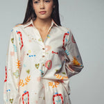 Picture of a woman wearing a white silk button up top with matching pants. Clothing set has bright shapes and plant and sun themed pictures. 