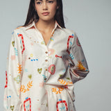 Picture of a woman wearing a white silk button up top with matching pants. Clothing set has bright shapes and plant and sun themed pictures. 