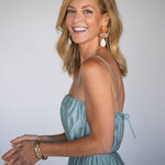 Woman wearing a light blue dress with a plain background. She is modeling a pair of large dangle statement earrings in neutral tones.