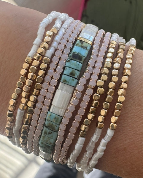 Picture of a woman's wrist displaying 11 bracelets. 6 bracelets are dainty white and gold beads, 4 bracelets are white quartz beads and the main bracelet is light mottled turquoise and mother of pearl beads.