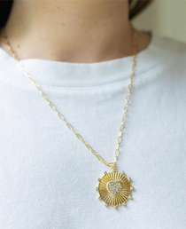 Girl wearing a necklace around her neck with a gold thin chain with a sun/heart medallion attached to it