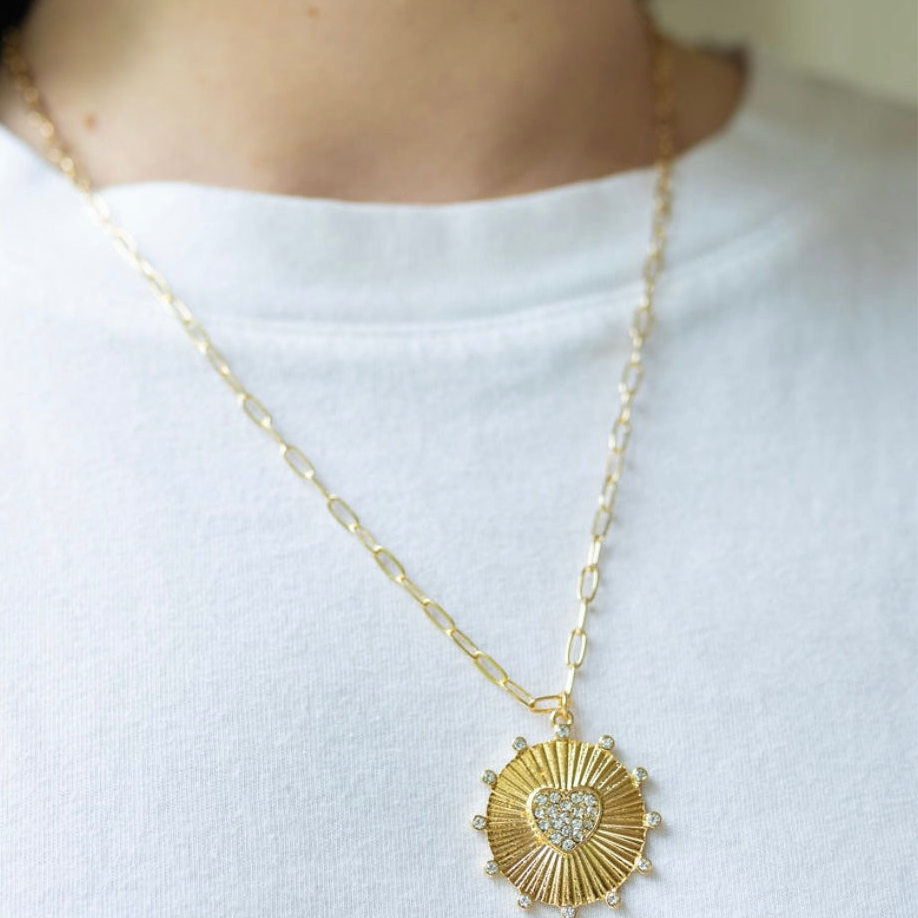 Girl wearing a necklace around her neck with a gold thin chain with a sun/heart medallion attached to it