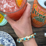Picture of a hand holding an iced glass of a pink drink and wearing a gold cuff style bracelet with inlaid turquoise beads.