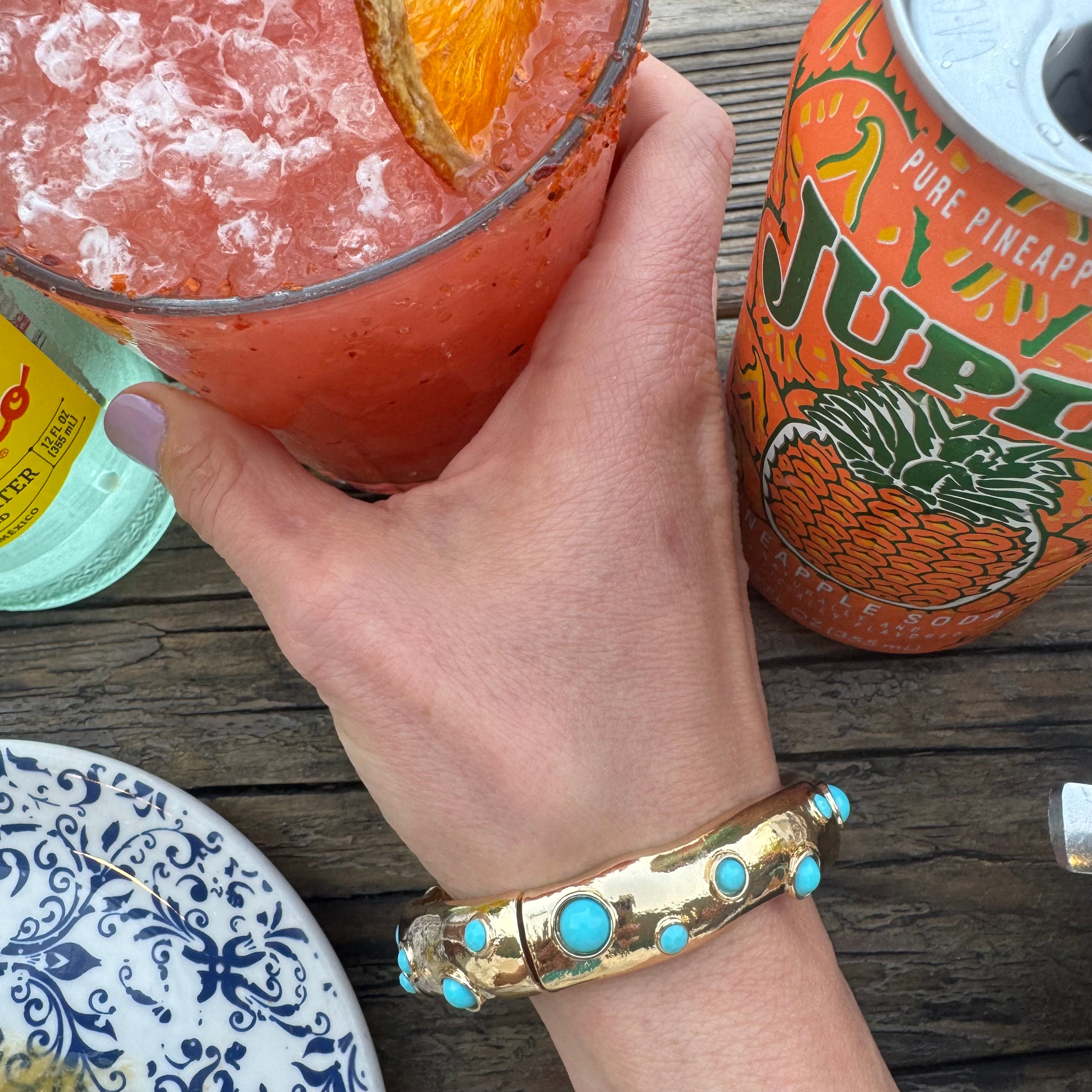 Picture of a hand holding an iced glass of a pink drink and wearing a gold cuff style bracelet with inlaid turquoise beads.