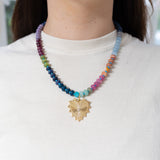Picture of model wearing a multi-colored beaded necklace of blues, pink, orange, green and purples. Necklace also has large gold heart pendant with a diamond in its center.