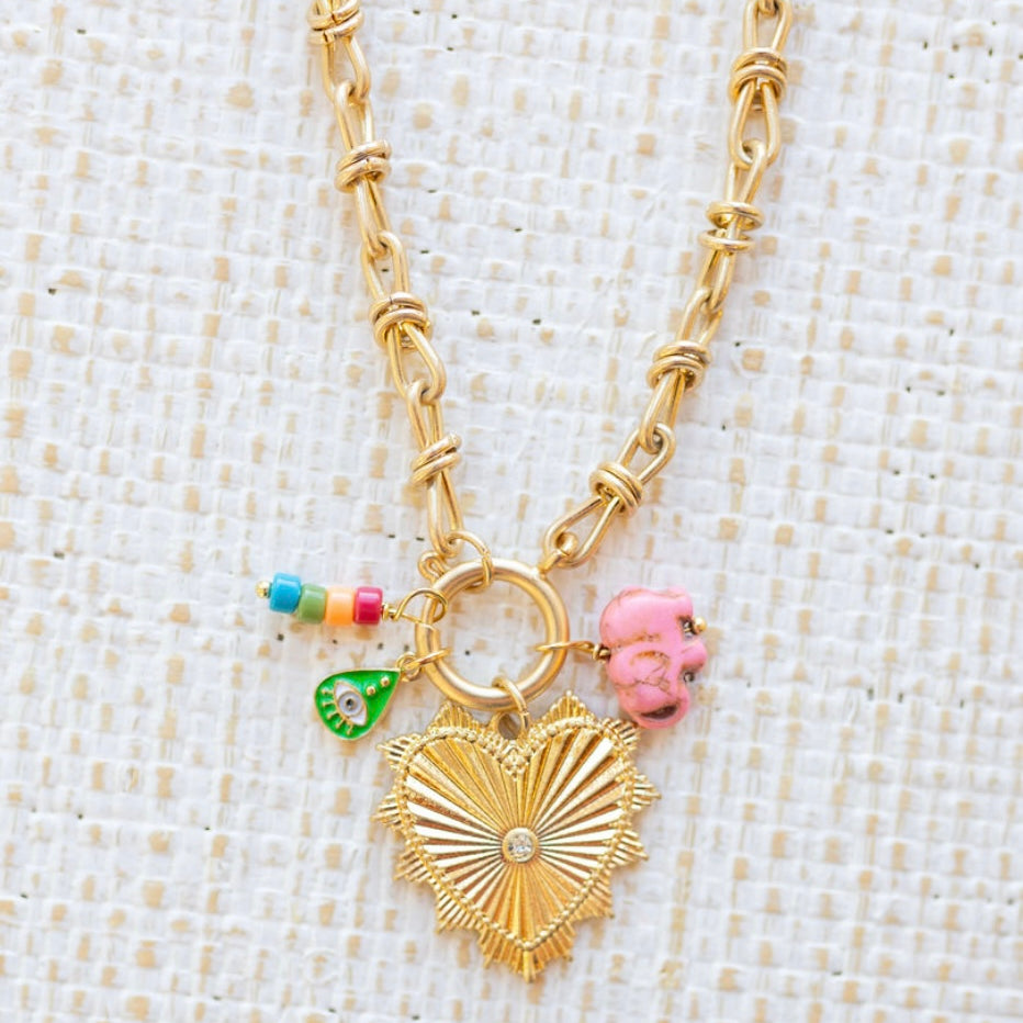 Gold necklace toggle chain with large gold heart pendant with a diamond center.  Three charms attached, one of several multicolored beads, a green evil eye and a pink elephant.