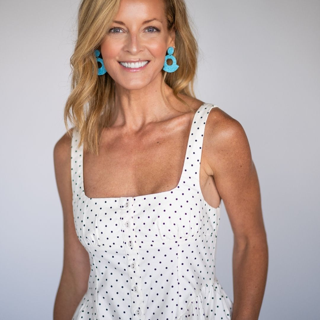 Woman wearing a white polka dot dress with blue statement earrings against a plain background